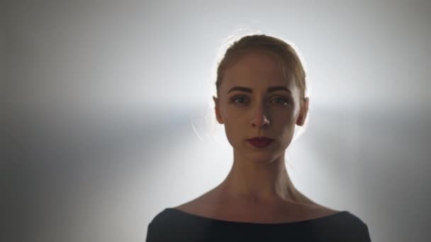 Portrait of confident gorgeous female ballet dancer standing in spotlight looking at camera and looking away. Beautiful elegant graceful Caucasian ballerina posing in backlit fog indoors. — Stockvideo