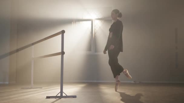 Широкий снимок уверенной худой балерины в пуантах, идущей на цыпочках к перекладине в подсвеченном тумане. Великолепная брюнетка в черном костюме репетирует в студии дымовых танцев. — стоковое видео