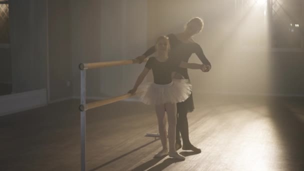 Smiling slim ballet teacher rehearsing tendu with student at barre in backlit fog. Wide shot of positive Caucasian woman and girl talking at ballet lesson in studio indoors. Happiness and art concept. — Vídeo de Stock