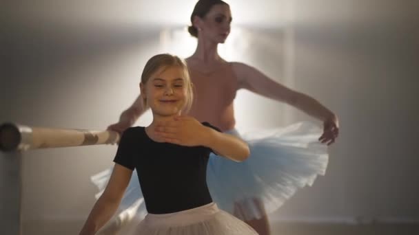 Smiling happy girl in tutu rehearsing third ballet position with blurred woman at background. Portrait of satisfied talented Caucasian student and professional teacher at barre in backlit fog. — Vídeo de Stock