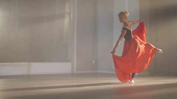 Wide shot of slim professional ballerina in arabesque ballet position holding scarlet red skirt bending. Graceful Caucasian female ballet dancer rehearsing performance in backlit fog on the right. — Wideo stockowe