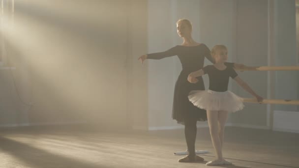 Gros plan d'un professeur de ballet souriant et d'un élève en troisième position répétant à droite dans un brouillard rétroéclairé. mince caucasien belle femme et mignon fille formation ballroom mouvement dans studio fumée. — Video