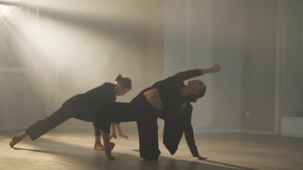 Zwei flexible grazile schlanke Balletttänzerinnen proben im hinterleuchteten Nebel im Studio drinnen. Charmante kaukasische Ballerinen in schwarzen Anzügen üben Performance im Scheinwerferlicht in Zeitlupe. — Stockvideo