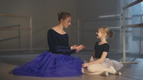 Weitwinkel von positiver Ballettlehrerin und Schülerin, die auf dem Boden im Tanzstudio sitzen, reden und in Zeitlupe lächeln. Anmutige Frau und Mädchen in Tutu und Pointen, die Pause beim Unterricht drinnen haben. — Stockvideo