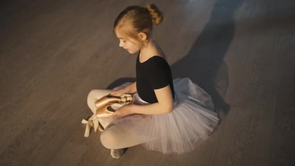 High angle view of smiling happy ballet student sitting on floor admiring new pointe shoes. Wide shot portrait of pretty charming Caucasian girl in tutu dreaming in dancing studio. Motivation concept. — Vídeo de Stock