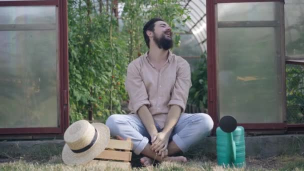 Satisfeito feliz sorrindo agricultor masculino sentado na entrada da estufa alongando-se olhando ao redor. Ampla foto de morena caucasiano barbudo homem com bigode descansando na pausa colheita de legumes. — Vídeo de Stock