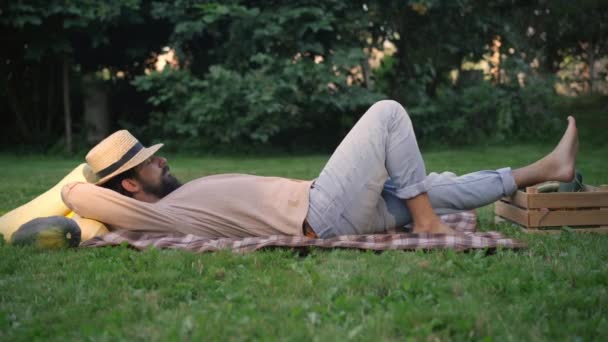 Side view of young male farmer sleeping lying on green meadow with harvest. Wide shot of tired Caucasian man taking a nap outdoors on sunny autumn day. Agriculture and harvesting concept. — ストック動画