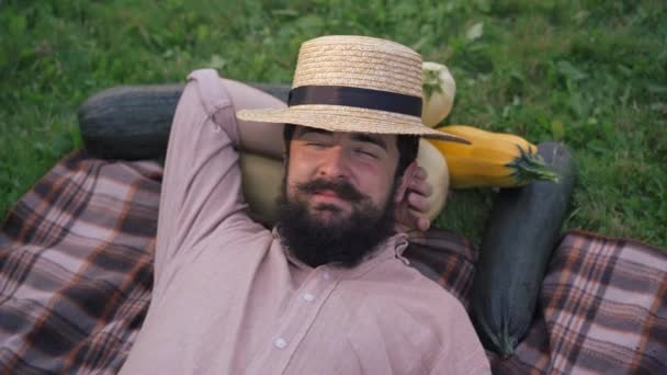 Vista superior de alegre joven granjero barbudo guapo acostado sobre una manta a la luz del sol con los ojos cerrados riendo. Hombre caucásico durmiendo relajado tomando una siesta cosechando cultivos al aire libre en un día soleado. — Vídeo de stock