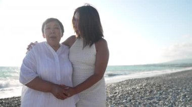 Gülümseyen güzel, yetişkin bir Asyalı kız ve neşeli yaşlı bir anne Akdeniz sahilinde şarkı söyleyerek dans ediyorlar. Turkuaz sahilde tatilin tadını çıkaran mutlu kadınların portresi.