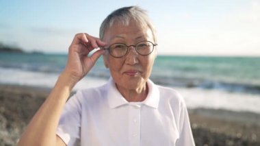 Akdeniz sahilinde yavaşça hareket eden turkuaz dalgalarla gülümseyen gözlüklü emekli Asyalı kadın portresi. Gün batımında poz veren kadın.