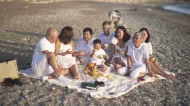 Kayalık deniz kıyısında battaniyeye oturmuş ağır çekimde kahkahalarla gülen Kafkas bir adamla mutlu bir Asyalı aile fotoğrafı. Rahat erkekler, kadınlar ve çocuklar tatil yerlerinde aile tatillerinin tadını çıkarıyorlar..