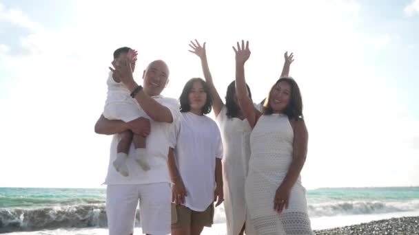 Família multigeracional asiática alegre acenando em câmera de câmera em pé à luz do sol no resort turístico mediterrâneo. Feliz homem mulheres e bebê menina posando em férias na praia do mar. — Vídeo de Stock