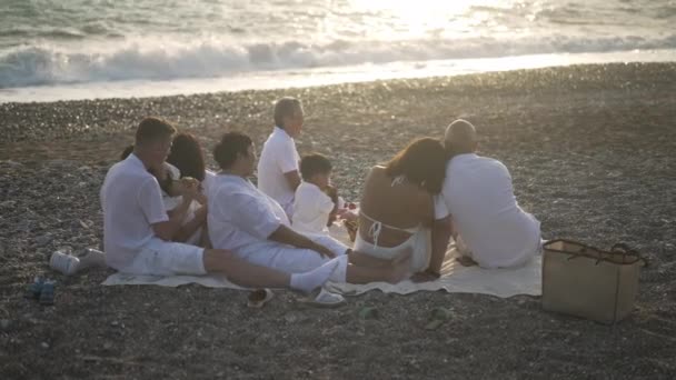 Wide shot back view of relaxed Asian family admiring sunset on Mediterranean sea coast with foamy waves splashing at background with sun reflecting in water. Tourism and leisure concept. — Stock Video