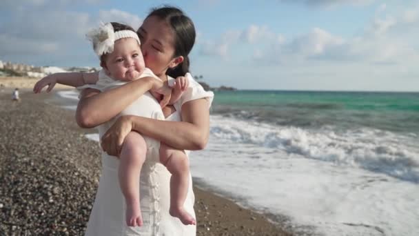 Charmante süße Baby-Mädchen mit asiatischer Mutter bei Sonnenuntergang an der Mittelmeerküste bewundern Schönheit der türkisfarbenen Wellen rollen am Strand in Zeitlupe. Porträt eines Kindes mit Eltern im subtropischen Resort. — Stockvideo