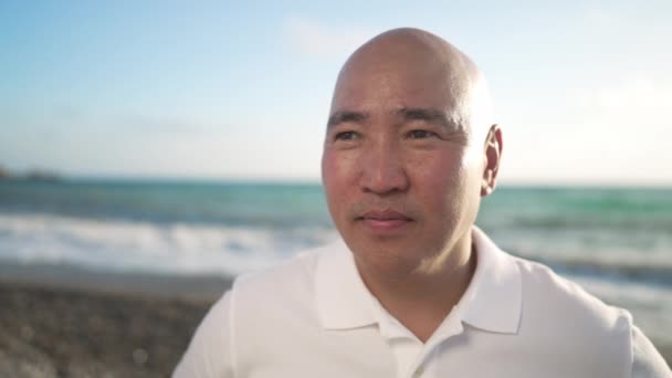 Hombre calvo positivo asiático de mediana edad mirando a la cámara sonriendo con olas turquesas del mar Mediterráneo rodando en la costa al fondo en cámara lenta. Retrato de turismo masculino feliz posando al atardecer. — Vídeos de Stock