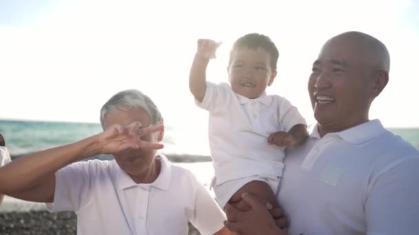 Vreugdevolle Aziatische overgrootvader en kleine jongen dansen in het zonlicht aan de Middellandse Zee. Positieve uitgebreide familie met plezier op het strand bij zonsondergang buiten. Gezinsvrijetijdsconcept. — Stockvideo