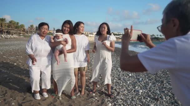 Brede opname van gelukkige multigenerationele Aziatische vrouwen die zingen in zonlicht aan de Middellandse Zee met wazige oma die video 's opneemt voor sociale media op de smartphone. Toerisme en roem. — Stockvideo