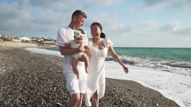 행복 한 젊은 백인 남자와 아시아 여자가 아름다운 지중해 해변을 따라 천천히 움직 이는 귀여운 여아와 함께 걷고 있는 모습. 해질 녘에 관광 휴양지에 있는 어린 부모와 딸. — 비디오