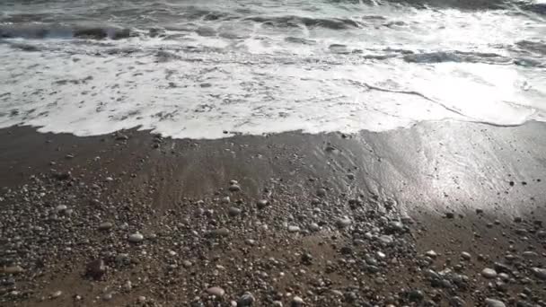 Vista de alto ângulo da espumosa água do mar do oceano rolando sobre a praia de areia com pequenas rochas em raios de sol ao ar livre. Ondas limpas do mar Mediterrâneo salpicando no raio de sol ao pôr do sol. Capa marinha pitoresca. — Vídeo de Stock