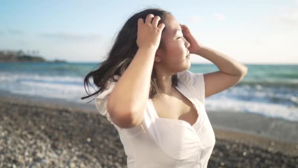 Eccitato felice giovane donna asiatica lanciando i capelli guardando la fotocamera sorridente come onde turchesi rotolando al rallentatore sullo sfondo. Ritratto di positivo bel millenario in posa sulla costa mediterranea. — Video Stock