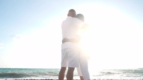 Weitwinkelaufnahme eines sich umarmenden asiatischen Mannes und einer Seniorin, die in der Sonne am Strand stehen, während türkisfarbene schäumende mediterrane Wellen in Zeitlupe über die Küste plätschern. Glücklicher Sohn und Mutter bewundern Sonnenuntergang im Resort — Stockvideo