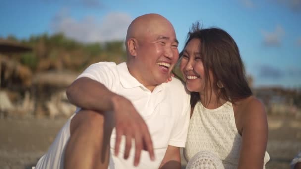 Aimant couple asiatique adulte assis à la lumière du soleil sur la mer Méditerranée pittoresque riant admiratif coucher de soleil. Portrait d'un homme et d'une femme d'âge moyen heureux profitant de vacances en famille à la station. — Video