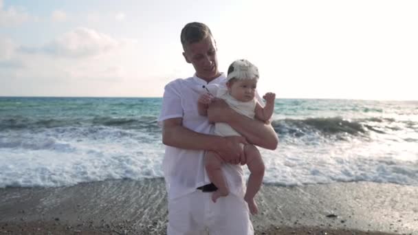 Der junge kaukasische Vater hält seine asiatische Tochter in den Händen und winkt lächelnd in die Kamera. Porträt eines positiven Mannes posiert mit einem kleinen Mädchen an der malerischen Mittelmeerküste bei Sonnenuntergang in Zeitlupe. — Stockvideo