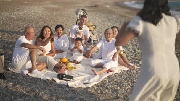 Rindo família asiática feliz e homem caucasiano adivinhando palavras como mulher gesticulando em câmera lenta. Ampla foto de pessoas multigeracionais positivas se divertindo sentadas na praia do mar Mediterrâneo ao pôr do sol. — Vídeo de Stock