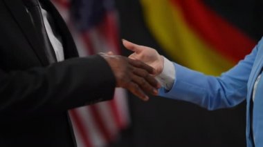 Ulusal bayrakların arkasında Afro-Amerikan erkek ve beyaz kadın el sıkışması. Tanımlanamayan diplomatlar basın toplantısında siyah arkaplanda el sıkışıyorlar.
