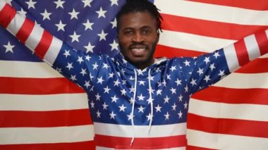 Mutlu Afro-Amerikan gururlu adam, Amerikan bayrağını tutarken ellerini uzatmış kameraya bakıp gülümsüyor. Amerikan bayrağı desenli kapüşonlu, ulusal sembollü neşeli ve kendinden emin bir adamın portresi..