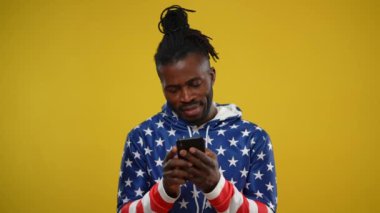 Happy, sosyal medya mesajlarında akıllı telefondan gülümseyen Afrikalı Amerikalı bir adamı özümsedi. Kapüşonlu, sarı arka planda Amerikan bayrağı izi olan cihaza bağımlı adamın portresi..