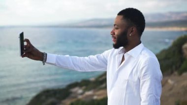 Mutlu Afro-Amerikan eşcinsel turistin akıllı telefonuyla günbatımının güzelliğini filme almasının yan görüntüsü. Gülümseyen yakışıklı LGBT adamı, sakin mavi Akdeniz koyunun arka planında duran doğaya hayran..