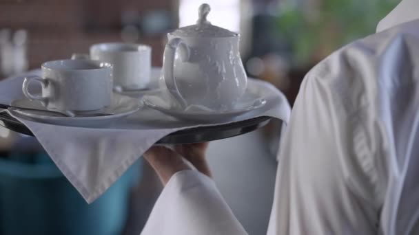 Close-up tray with teapot and white tea cups in male Caucasian hand. Unrecognizable man waiter walking in slow motion carrying order for guests in luxurious restaurant. — Stock Video