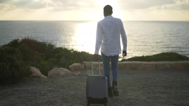 Επιστροφή άποψη της Αφρικής Αμερικανός άνθρωπος με αποσκευές σε τσάντα τρόλεϊ με τα πόδια σε αργή κίνηση προς το ηλιοβασίλεμα στη Μεσόγειο θάλασσα θέρετρο. Ευρεία βολή της ευτυχούς αρσενικό τουρίστα με αποσκευές θαυμάζοντας την ομορφιά του θαλασσινού τοπίου. — Αρχείο Βίντεο
