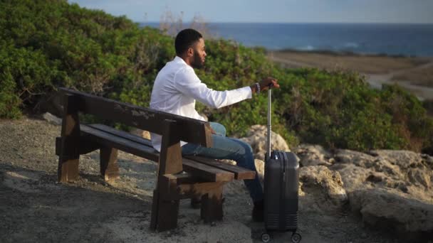 Široký pohled na Afroameričana, jak sedí na lavičce s taškou na vozíky a dívá se na tyrkysové Středozemní moře. Důkladné LGBT osoba se zavazadly v turistickém letovisku těší klid. — Stock video