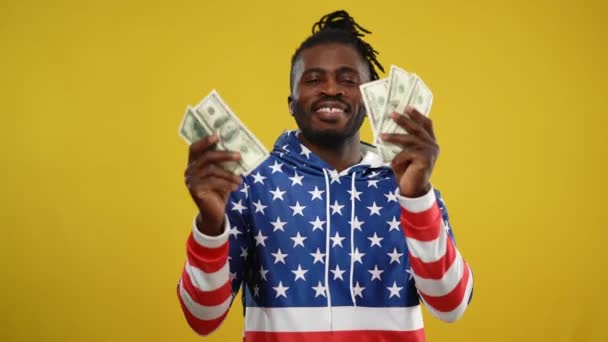 Emocionado rico afroamericano sonriendo hombre bailando en el fondo amarillo sonriendo. Retrato de tiro medio de feliz hipster rico y exitoso con capucha con estampado de bandera estadounidense posando con dinero. Presumiendo. — Vídeos de Stock