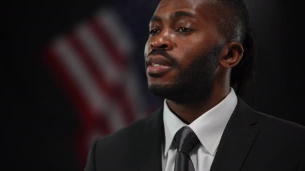 Retrato de close-up de diplomata político afro-americano confiante respondendo a perguntas na conferência de imprensa. Um homem bonito e sério a falar com a bandeira americana no fundo preto. Fala em público. — Vídeo de Stock