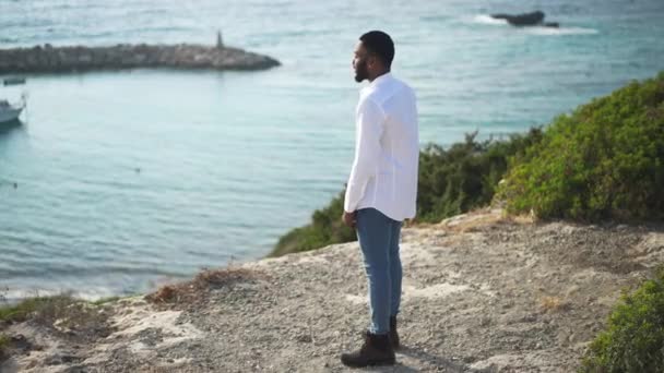 Ampla foto de rico relaxado homem afro-americano feliz de pé ao pôr do sol na costa do mar Mediterrâneo admirando iate borrado na baía. Sorrindo jovem bonito cara descansando em férias. — Vídeo de Stock