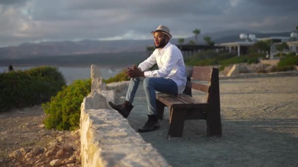 Vista lateral de homem gay afro-americano pensativo sentado em raios de sol no banco olhando para longe. Romântico LGBT pessoa descansando na costa do mar Mediterrâneo com paisagem pitoresca no fundo. — Vídeo de Stock