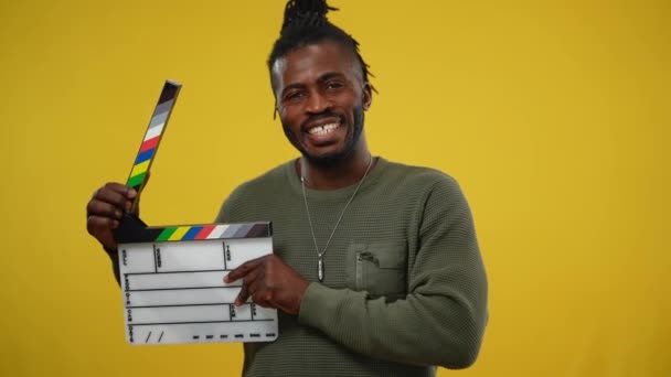 Afro-americano seconda telecamera assistente cliccando clapperboard su sfondo giallo. Ritratto di uomo bello allegro sorridente che posa guardando la macchina fotografica. Produzione cinematografica e video. — Video Stock
