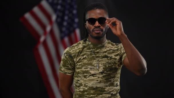 Portrait of confident African American military man in khaki T-shirt taking off sunglasses crossing hands looking at camera. Medium shot of proud recruit posing at black background with USA flag. — Stock Video