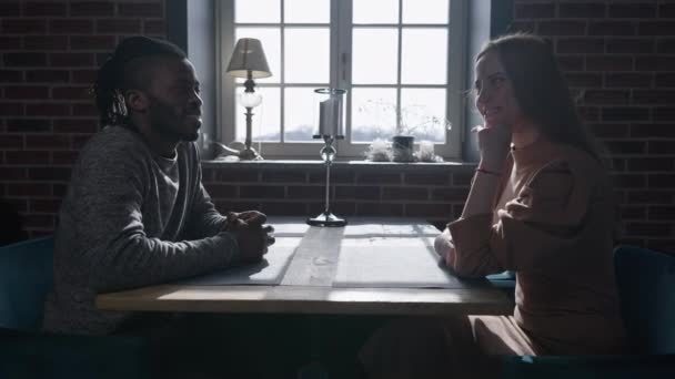 Side view of happy loving Afro-Amerikaanse man en blanke vrouw zitten aan tafel in restaurant praten glimlachend. Jong stel dat uitgaat in café binnen. Relatie en vrijetijdsconcept. Langzame beweging. — Stockvideo