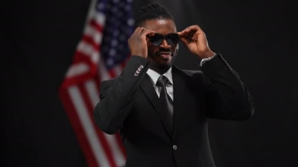 Портрет щасливого афроамериканського чоловіка в костюмі, який одягає сонцезахисні окуляри, танцює і посміхається, коли в камері спалахують долари. Радісний чоловік - політик, який радіє виборам на чорному фоні. — стокове відео