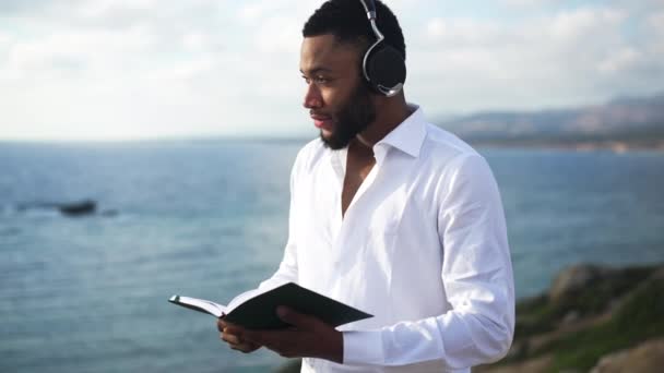 Przystojny Afroamerykanin czyta książkę na wybrzeżu Morza Śródziemnego o wschodzie słońca patrząc na krajobraz morski. Portret pewnej siebie inteligentnej osoby LGBT cieszącej się hobby na świeżym powietrzu. — Wideo stockowe