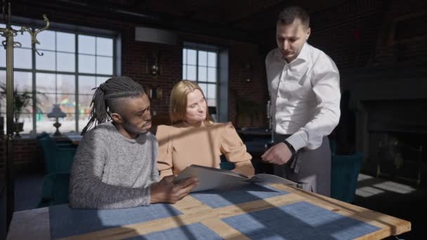 Camarero ayudando a los huéspedes a elegir el almuerzo en un lujoso restaurante sentado a la mesa a la luz del sol. Caucásico hombre hablando con interracial pareja citas comedor en el interior. Movimiento lento. — Vídeo de stock