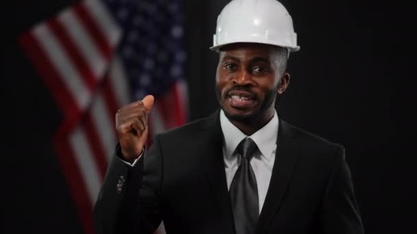 身穿正装、头戴硬礼帽的快乐英俊的非洲裔美国男人，在展示房地产开发项目时，微笑着竖起大拇指看着镜头。有信心的政治家竞选. — 图库视频影像