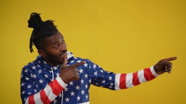 Retrato de homem afro-americano alegre sorrindo apontando para a direita no fundo amarelo. alegre relaxado positivo cara no hoodie com bandeira dos EUA impressão posando mostrando anúncio. — Vídeo de Stock