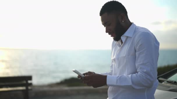 Homem LGBT confiante navegando na Internet no smartphone ao pôr do sol na costa do mar Mediterrâneo. médio tiro de bonito bem sucedido afro-americano gay empresário planejamento startup ao ar livre admirando natureza. — Vídeo de Stock