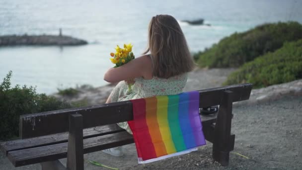 Baksidan bild av vackra unga kaukasiska brunett kvinna sitter på bänk med hbt flagga innehav bukett gula tulpaner beundrar solnedgången på Medelhavet. Avslappnad glad turist njuter av fritid. — Stockvideo
