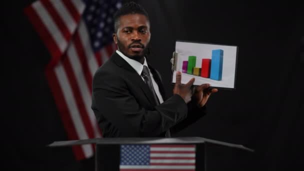 Schöner afroamerikanischer Mann, der auf ein Diagramm zeigt, das mit der Flagge der USA vor schwarzem Hintergrund spricht. Porträt eines selbstbewussten, ernsthaften männlichen Politikers Diplomat, der Öffentlichkeit und Journalisten über Erfolge berichtet. — Stockvideo